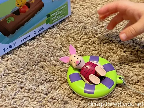 A plastic Piglet from Winnie The Pooh in a green and purple raft in front of a box and a little kid hand going to grab Piglet.