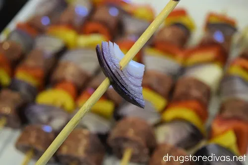 A piece of red onion on a skewer, hovering over completed sausage kabobs.