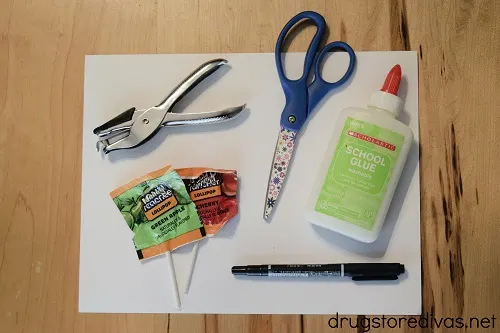 A hole punch, scissors, glue, black marker, lollipops, and white card stock on a table.