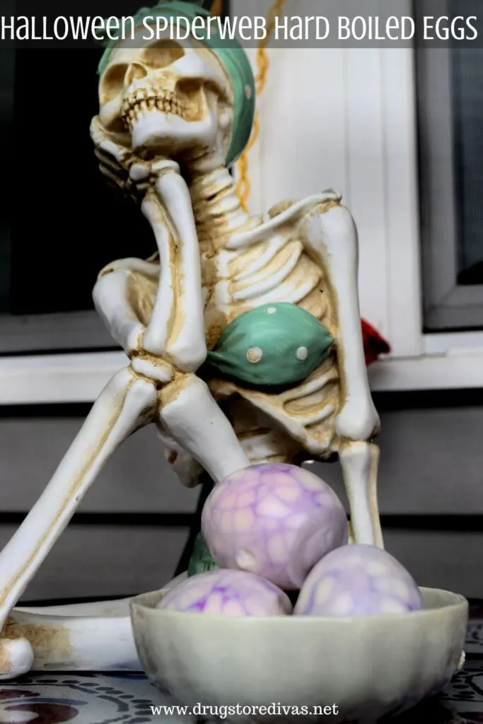 A bowl of hard boiled eggs with spiderwebs designs on them in front of a skeleton Halloween decoration with the words "Halloween Spiderweb Hard Boiled Eggs" digitally written on top.