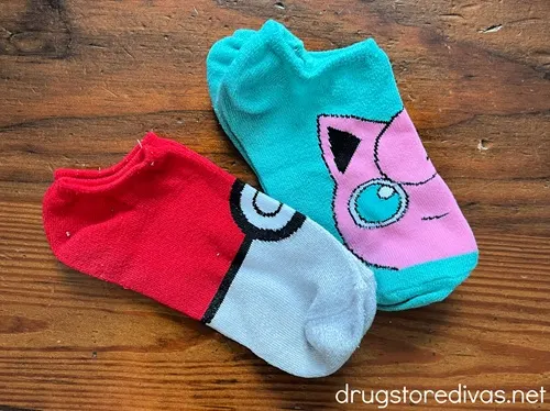 Two pairs of Pokemon themed socks (one is a pokeball and one is teal with a pink character on it) on a table.