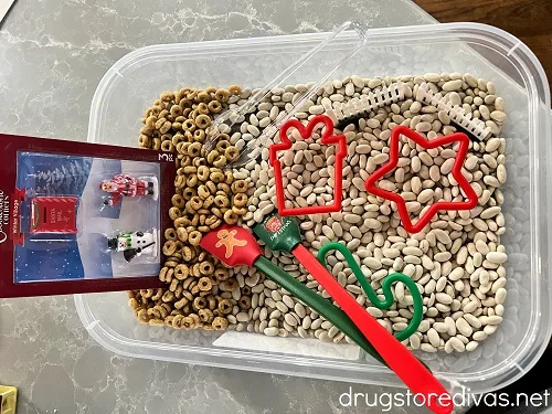 A pair of tongs, Christmas cookie cutters, spatulas, and figurines in a plastic bin with Cheerios and beans.