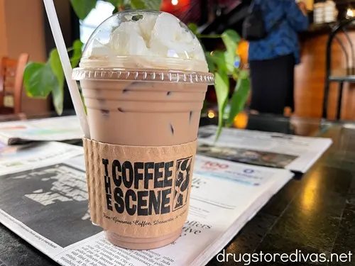 An iced coffee with whipped cream on a newspaper in a coffee shop with a label that says The Coffee Scene on it.