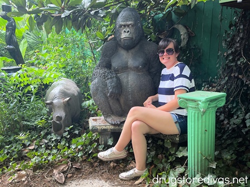 A woman in a blue and white striped shirt, shorts, and sunglasses sitting on a bench next to a gorilla statue that's next to another animal statue.