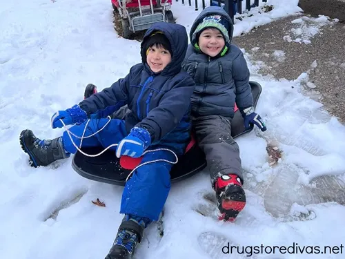Two boys wearing jackets, snow pants, boots, and hats on a sled in the snow.