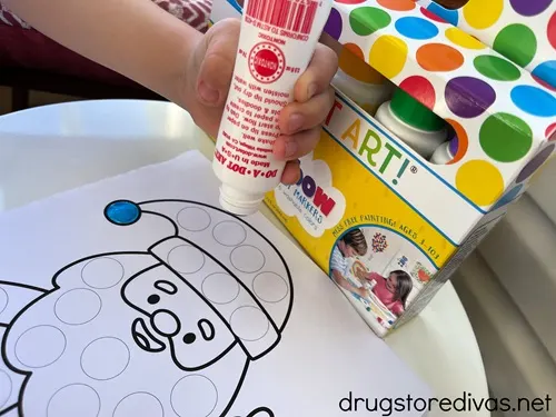 A hand holding a dot marker with a pack behind it dabbing a Santa coloring page.