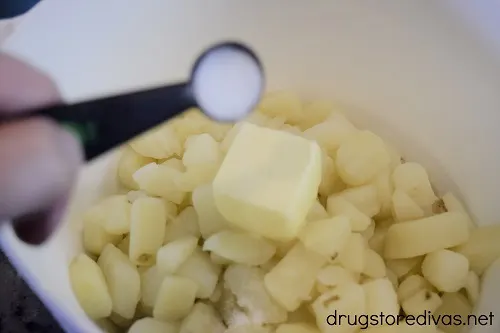 Salt being poured on top of a pat of butter on top of diced potatoes in a bowl.
