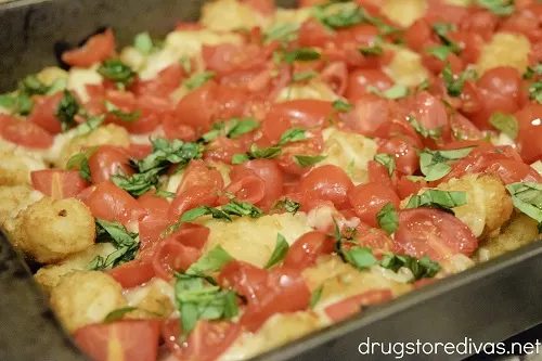 Basil, cherry tomatoes, and mozzarella cheese on top of tater tots in a cake pan.