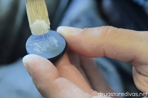 Two fingers holding a blue wooden disc and a paint brush painting white glue onto it.