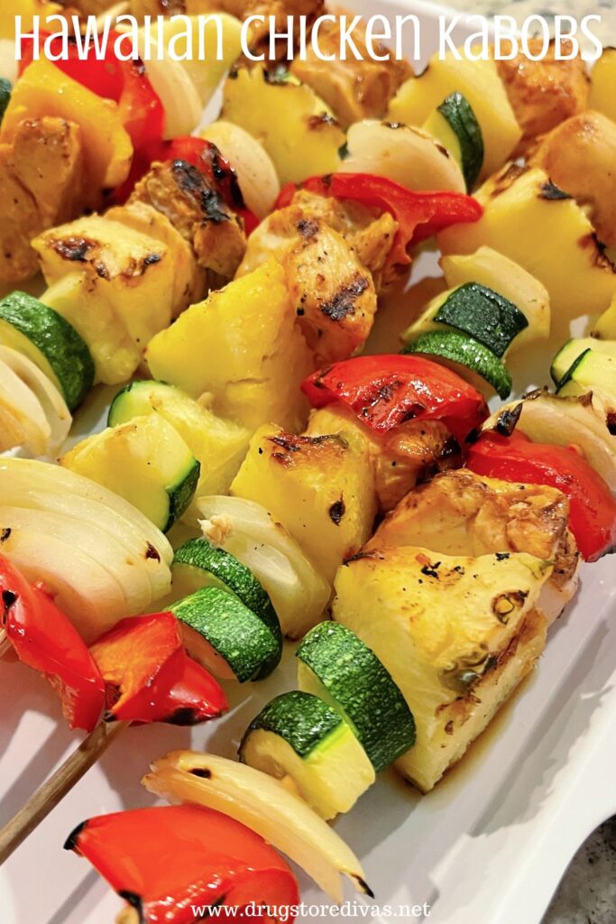 A bunch of skewers with vegetables and chicken on a white tray with the words "Hawaiian Chicken Kabobs" digitally written above them.