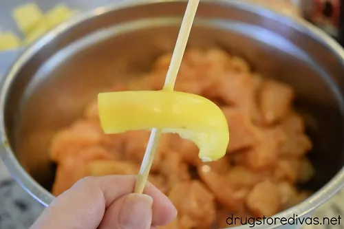 A hand holding a skewer with a piece of yellow pepper on it over a bowl of chicken.