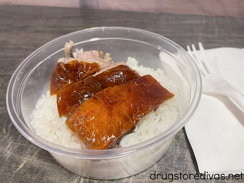 Pieces of roasted duck on top of rice in a clear plastic container next to a plastic fork on top of a napkin.