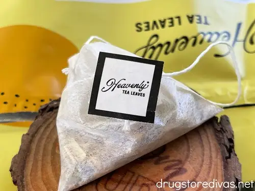 A tea bag with a tag that says Heavenly Tea on it on a wooden coaster with a yellow bag behind it.