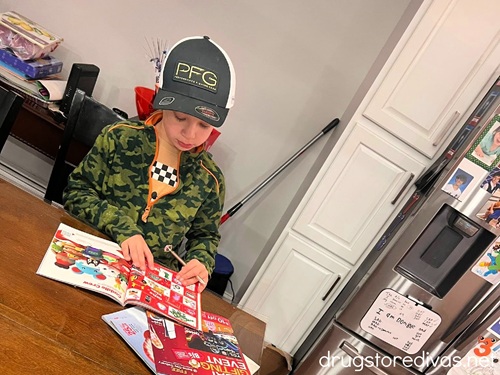 A little boy with a hat on reading magazines in a kitchen.