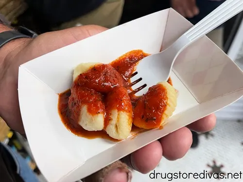 A hand holding a small paper tray with gnocchi and red sauce on top with a fork in it.