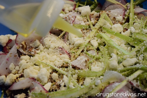 Olive oil being poured over shaved asparagus, shaved radish, pecans, and feta in a blue bowl.