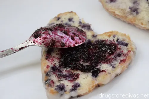 A heart-shaped blueberry biscuit with homemade blueberry jam spread on it and a spoon with jam resting on it.