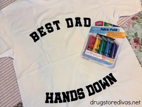 A white shirt that says "Best Dad Hands Down" on it with a pack of fabric paint next to it.