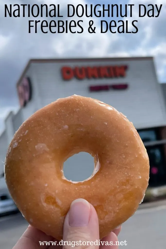 A hand holding a glazed doughnut in front of a doughnut shop with the words "National Doughnut Day Freebies & Deals" digitally written above it.