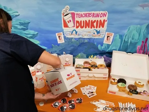 A woman pouring coffee on a table filled with donuts and a sign that says "Teachers Run On Dunkin" on the wall near her.