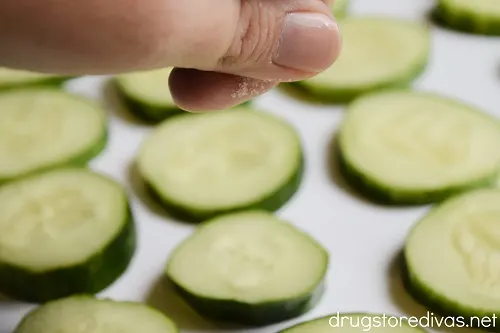 A hand sprinkling salt over cucumber slices on a white tray.