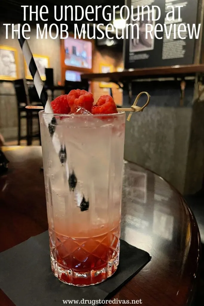 A cocktail with a black and white straw and raspberries on a shelf with a bar and displays behind it and the words "The Underground At The Mob Museum Review" digitally written on top.