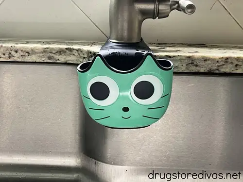 A teal cat face dish rag holder hanging off a faucet in a kitchen sink.