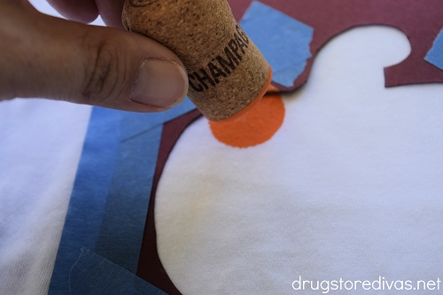 A wine cork with orange paint dabbing onto a pumpkin-shaped stencil on a shirt.