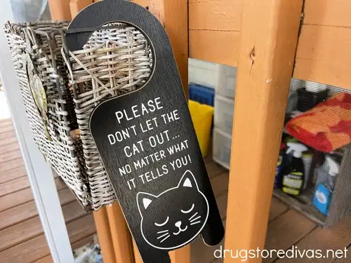 A black door hanger, shaped like a cat, that says "Please Don't Let The Cat Out No Matter What He Tells You" on it.