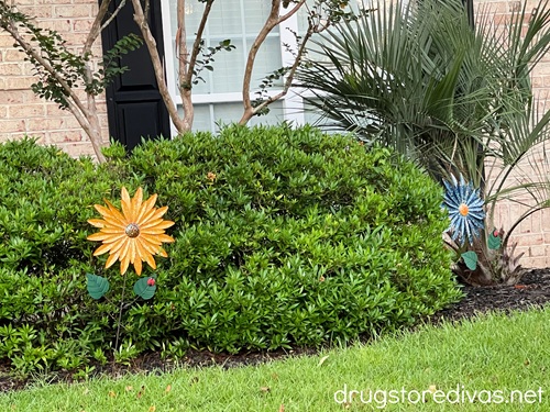 Two metal flowers, one yellow and one blue, on either side of a bush in front of a home.