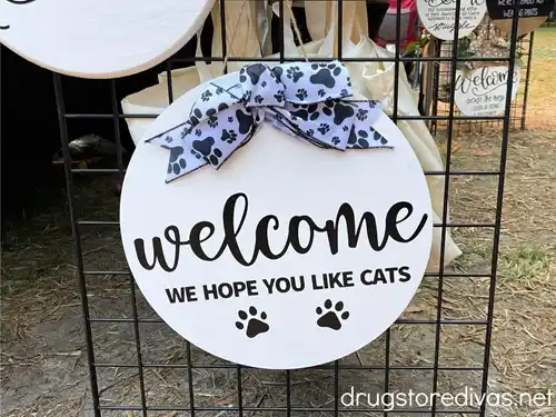 A white, round front door sign that says Welcome We Hope You Like Cats on it.
