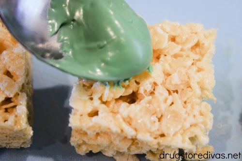 Melted green candy melts on a spoon being poured onto a Rice Krispy treat.