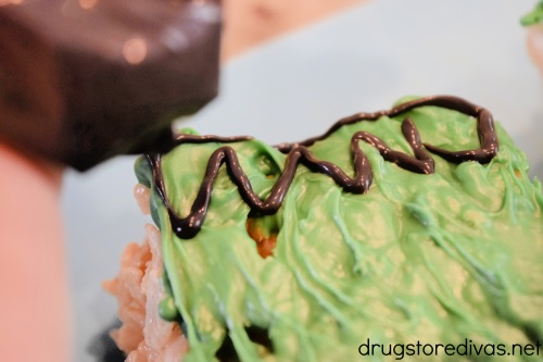 A hand holding a piping bag of black candy melts being used to pipe hair onto a green chocolate topped Rice Krispy Treat.