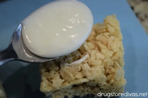 A spoon with white chocolate on it being poured onto a Rice Krispies Treat.
