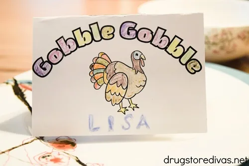 A place card with the words gobble gobble over a colored turkey and the name LISA at the bottom on top of plates.