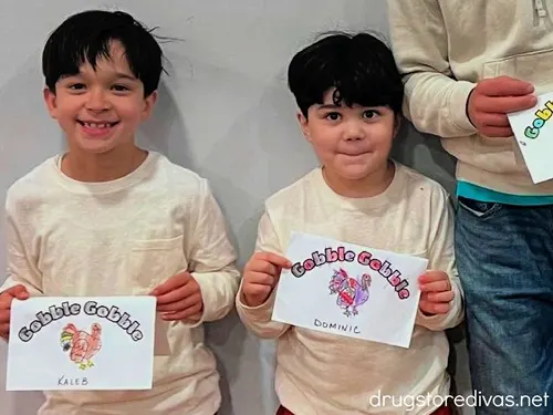 Two boys in beige tops holding Thanksgiving place cards that they colored.