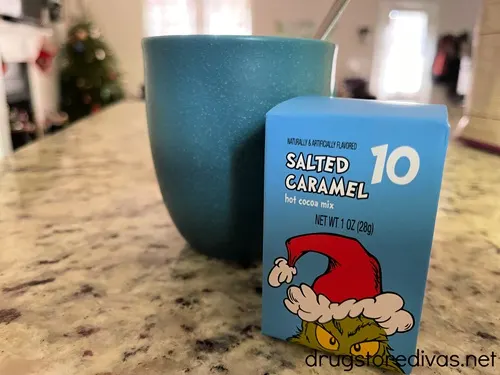 A blue box of hot chocolate with the Grinch on it in front of a blue cup with a spoon it in on a counter.