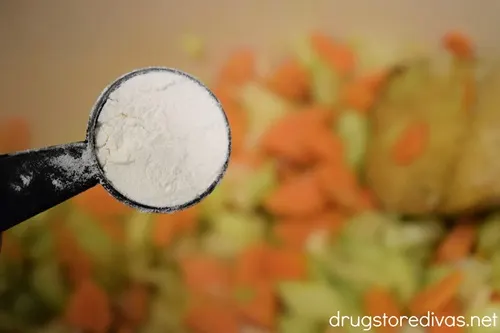 Flour in a measuring spoon above a Dutch oven with carrots and celery.