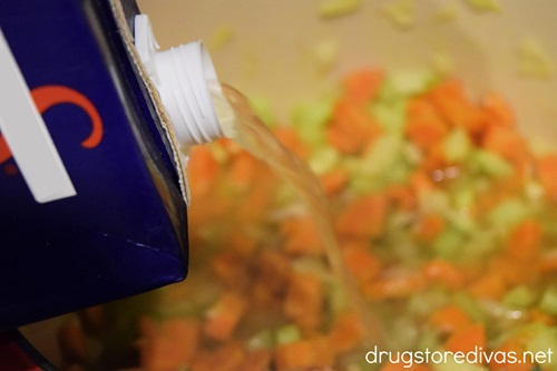 A box of chicken broth being poured on chopped carrots and celery.