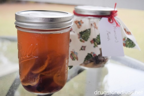 A mason jar filled with a fark liquid and fruit in front of a mason jar wrapped with fabric on top.