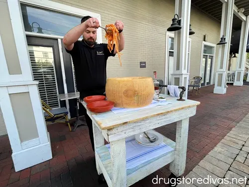 A man in a black shirt holding a serving of spaghetti in the air over two red bowls and a large parmesan cheese wheel that's on a white table. This is happening outside Tarantelli's restaurant in Wilmington, NC.