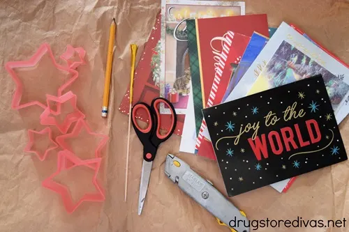 Six pink star shaped cookie cutters, a pencil, a foil pick, scissors, Christmas cards, and an X-Acto knife.
