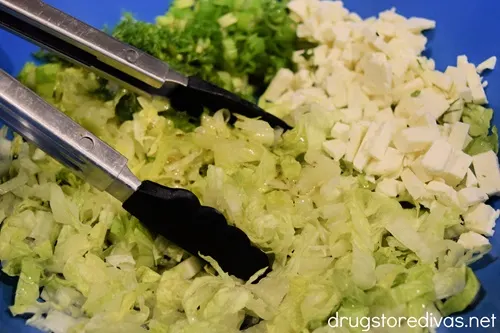 Tongs tossing a salad of chopped lettuce, green onion, feta cheese, and dill.