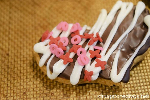 Pink and red XO-shaped sprinkles and white zigzag lines on a chocolate-covered heart shaped cookie.