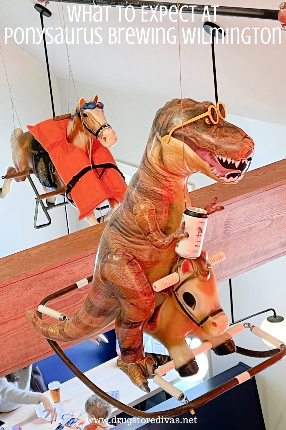 A carousel horse wearing a life preserver and an inflatable T-Rex holding a beer on a children's ride on toy horse, both suspended from the ceiling, with the words 