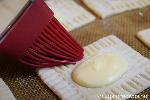 A pastry brush brushing the outside of of a rectangle of puff pastry that's filled with cream.