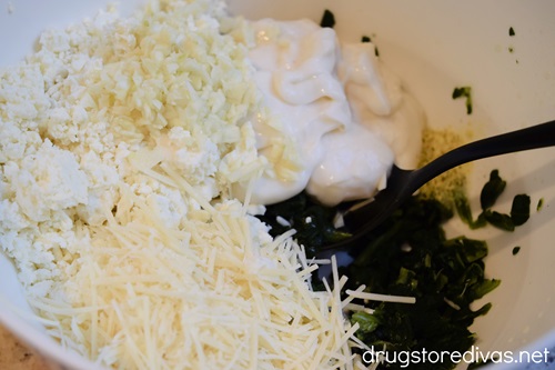 Feta cheese, parmesan cheese, chopped garlic, mayo, and spinach in a white bowl with a mixing spoon.