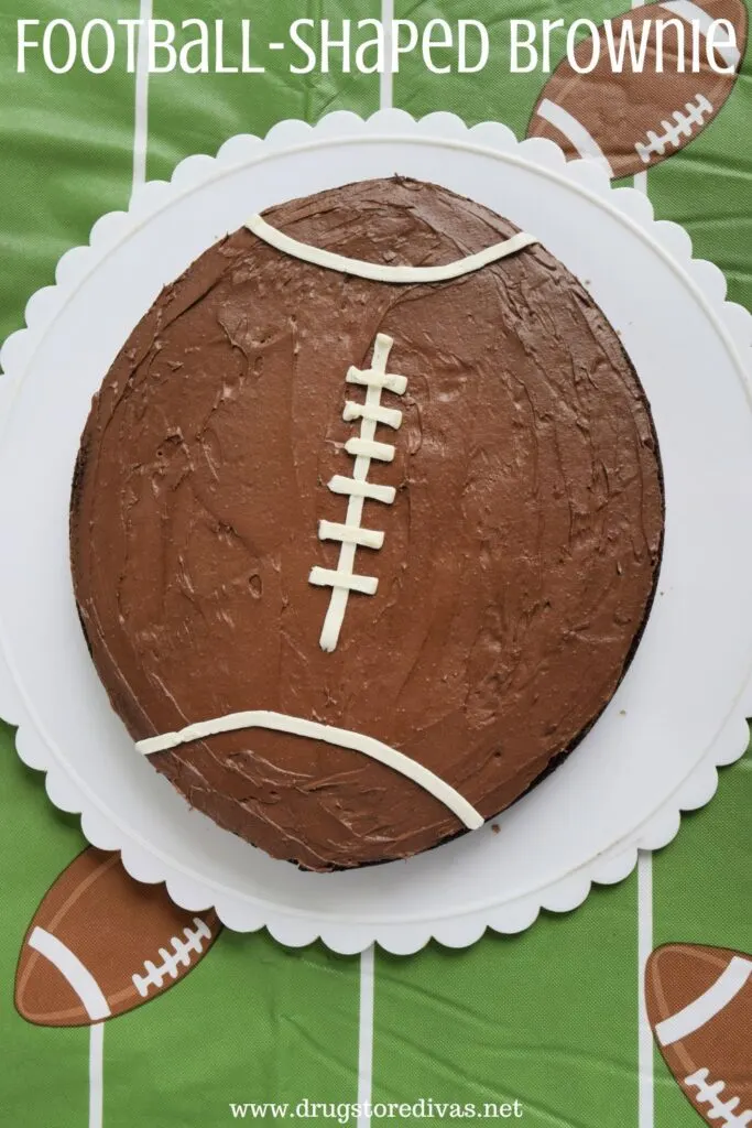 A brownie decorated like a football on a white plate with scalloped edges on a football-themed tablecloth with the words "Football-Shaped Brownie" digitally written on top.