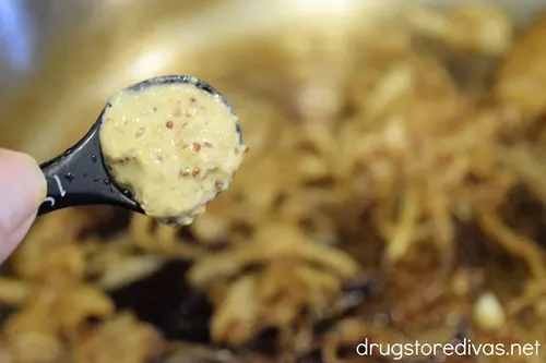 Mustard in a measuring spoon above sliced onions in a pan.