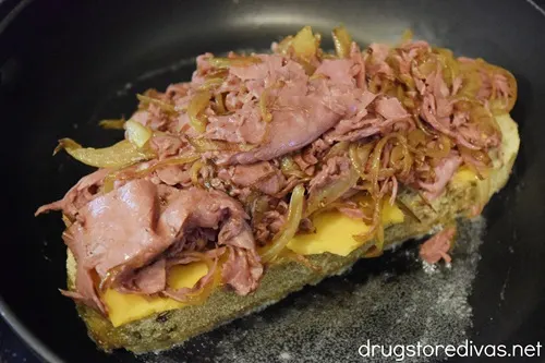 A slice of rye bread with cheese and sliced corned beef with onions on it on top of melted butter in a pan.